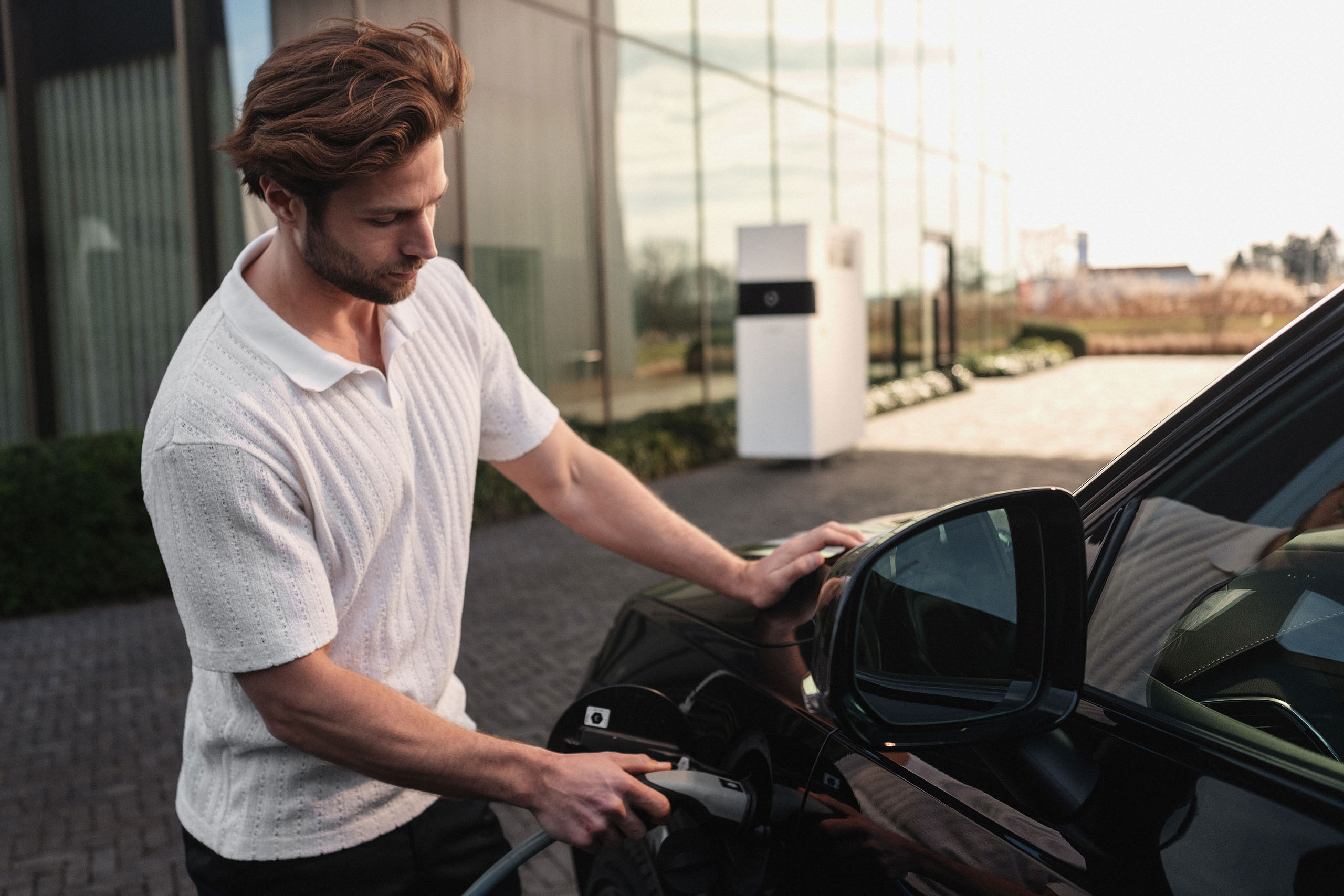 Man steekt de laadkabel in een elektrische auto, met een Kiwatt batterij op de achtergrond.